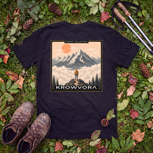 A black graphic 100% cotton hiking T-shirt laid flat on grass surrounded by pinecones, leaves, hiking boots, and trekking poles. The shirt features a large rectangular mountain landscape print with the text 'SUMMIT COLLECTION' at the top and 'KROWVORA' at the bottom.