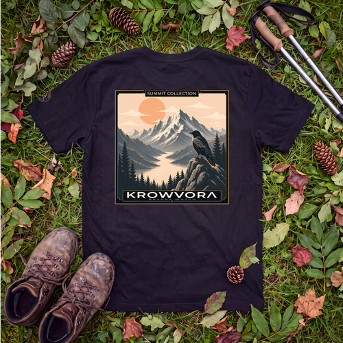 A black graphic 100% cotton hiking t-shirt laid flat on a bed of green grass, pine needles, pinecones, and hiking boots, with hiking poles in the top right corner. The graphic features a mountain landscape with a bird and the text 'SUMMIT COLLECTION' and 'KROWVORA'.